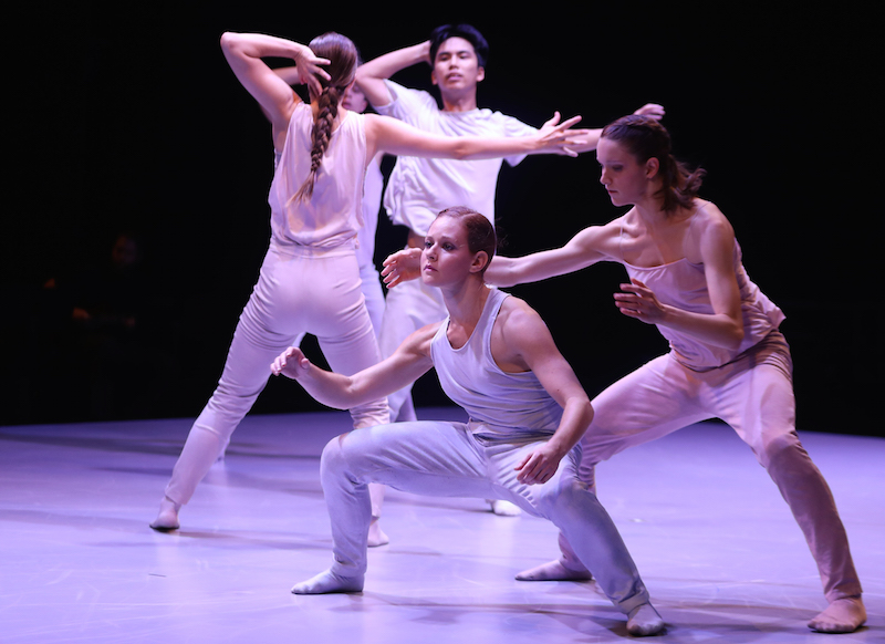 A women in white pants and t-shirt crouches; her legs are turned out and bent deeply at the knees. Other dancers in white are in the background. 